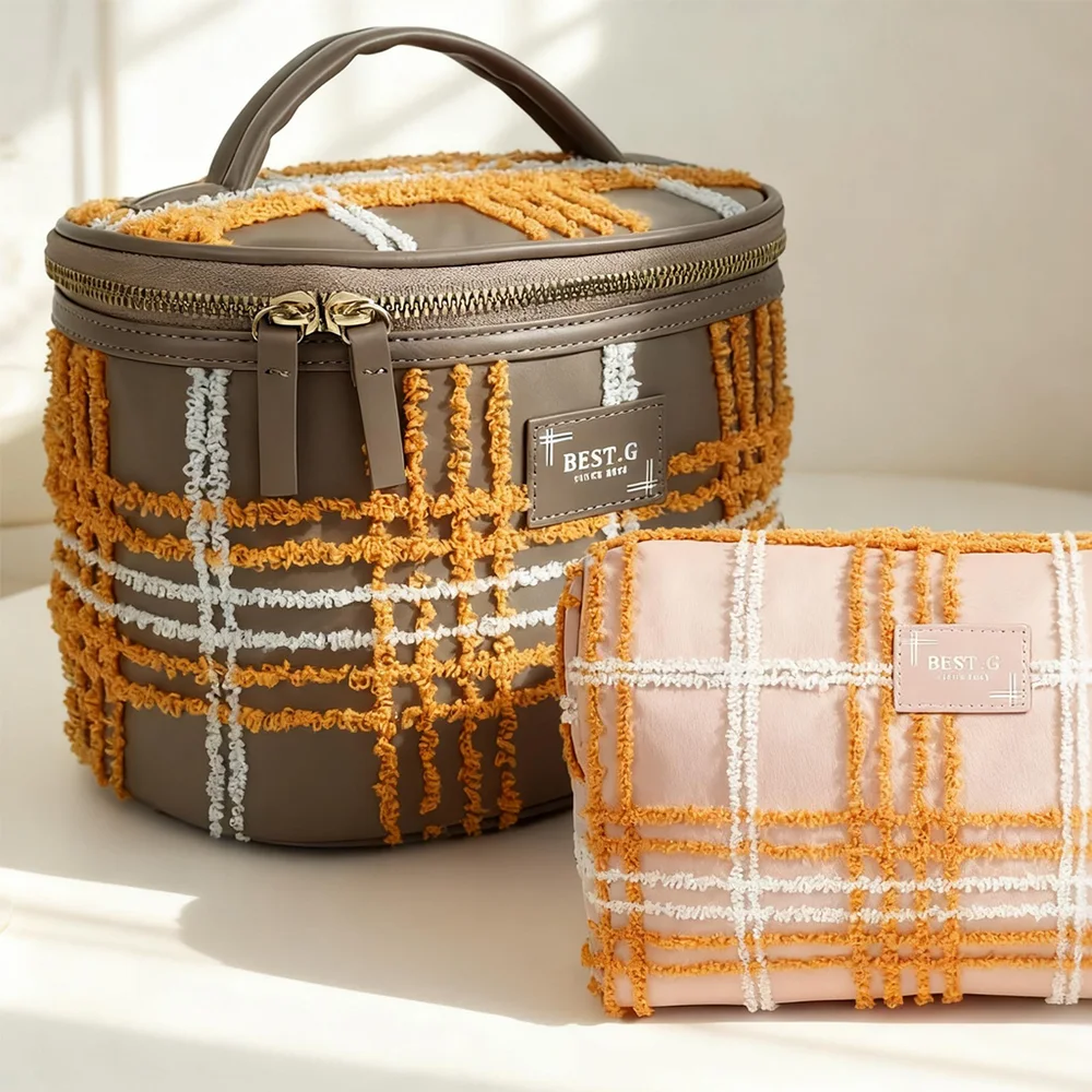 Taupe oval barrel cosmetic bag with mustard chenille plaid, gold zipper and top handle, beside blush-pink plaid pouch, on sunlit beige background